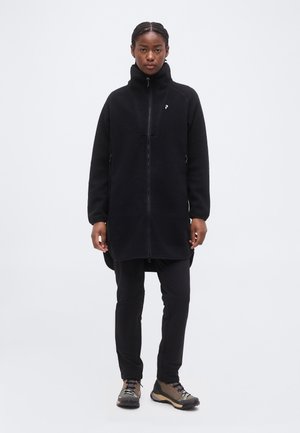 Γυναίκα με πλεξούδες, που φορά μακρύ μαύρο φλις μπουφάν, μαύρο παντελόνι και καφέ παπούτσια πεζοπορίας, στέκεται μπροστά σε απλό φόντο.