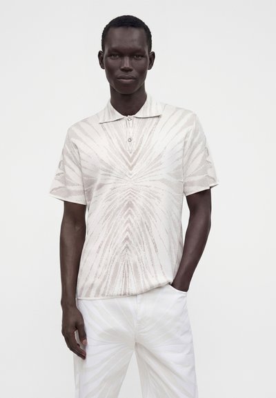 Homme portant un polo blanc à manches courtes à motifs et un pantalon blanc, une main dans la poche, debout devant un fond uni.
