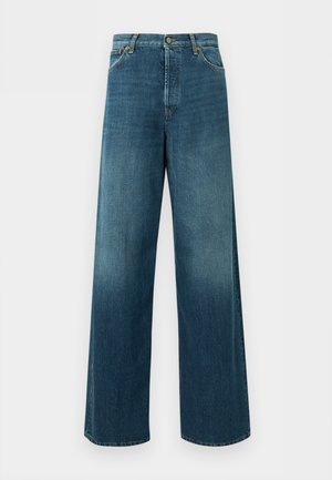 Femme portant un haut blanc ajusté à manches longues, un jean bleu à jambes larges avec les mains dans les poches et des sandales noires à bout ouvert, debout devant un fond uni.