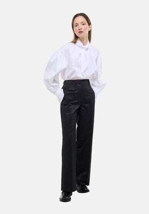 Femme debout les mains dans les poches, portant un chemisier blanc avec un nœud au col et un pantalon à taille haute noir à larges jambes, ainsi que des chaussures noires.