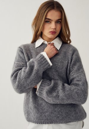 Giovane donna con capelli castani lunghi che indossa un maglione grigio lavorato a maglia sopra una camicia bianca con colletto, posando con una mano vicino al collo.