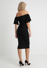 Robe noire décolletée épaules nues avec manches courtes volantes, silhouette ajustée et ourlet à hauteur des genoux. Portée avec des chaussures à talons noirs.