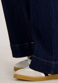 Donkerblauwe denim wijde broek met zichtbare stiksels, gecombineerd met lichtgrijze sneakers met witte accenten en een tan rubberen zool.