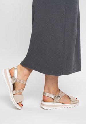 Feet wearing beige and metallic gold open-toe sandals with white soles, paired with a dark gray long skirt on a light background.