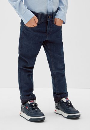 Enfant portant un jean bleu foncé, une chemise bleu clair et des baskets montantes bleu marine avec des semelles blanches, debout les mains dans les poches.