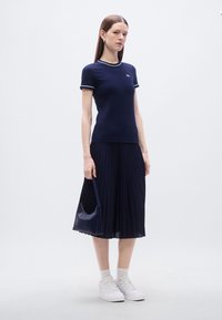 Μπλε σκούρο μπλουζάκι πόλο με κοντά μανίκια και λευκή περιμετρική λεπτομέρεια, συνδυασμένο με πλισέ μπλε σκούρη φούστα. Φορά λευκές κάλτσες και λευκά αθλητικά παπούτσια. Κρατά μικρή τσάντα στο χέρι.