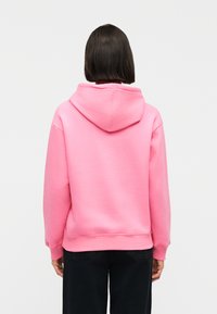 Άνθρωπος με κοντά σκούρα μαλλιά, ντυμένος με μονόχρωμο ροζ φούτερ με κουκούλα και μαύρο παντελόνι, εμφανίζεται από πίσω σε λευκό φόντο.