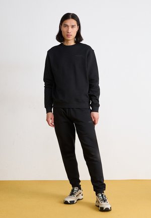Sort sweatshirt og matchende bukser lavet af blødt stof. Har en rund halsudskæring og ribbede manchetter. Bæres med beige træningssko.