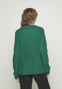 Donna con capelli mossi che indossa un maglione lungo a maniche lunghe verde testurizzato e pantaloni neri, di spalle su uno sfondo neutro.