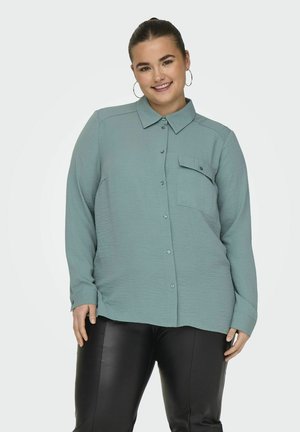 Chemise longue à manches longues turquoise clair avec un col, des boutons et une poche poitrine, associée à un pantalon en cuir synthétique noir. Tissu doux et texturé.