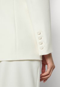 Gros plan sur une manche de costume blanche avec quatre boutons assortis et une main détendue sur le côté, sur un fond uni.