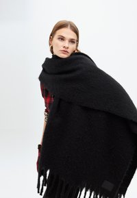 Černý šál s jemnou texturou, připomínající kožešinu, s třásněmi na okrajích a obdélníkovým tvarem. Na jedné straně má malou ozdobnou visačku.