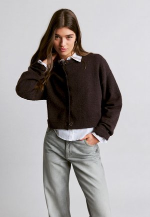 Young woman with long brown hair wears a dark brown buttoned cardigan over a white shirt and light gray jeans, standing against plain background.