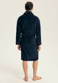 Mężczyzna stojący boso w ciemnoniebieskim, pluszowym szlafroku z paskiem i czerwonym wykończeniem, pokazany od tyłu na jednolitym tle.