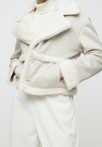Chaqueta de piel sintética beige con cuello y puños texturizados, que presenta botones de doble botonadura y bolsillos frontales. Material exterior suave.