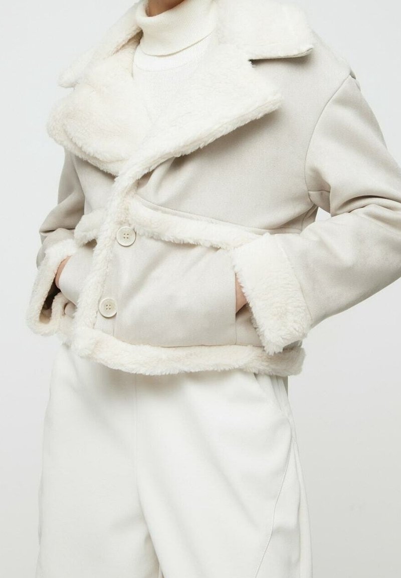 Chaqueta de piel sintética beige con cuello y puños texturizados, que presenta botones de doble botonadura y bolsillos frontales. Material exterior suave.