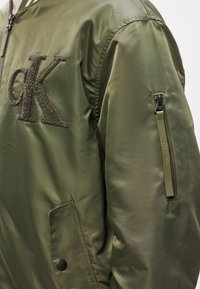 Oliiviroheline bomber-jakk musta tekstuurse "CK" logoga, varrukal tõmblukuga tasku ja siledalt läikiva viimistlusega.