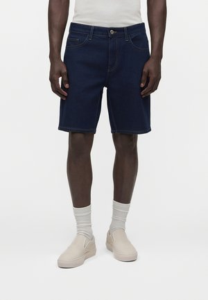 Mężczyzna w ciemnoniebieskich szortach jeansowych, białych skarpetkach, beżowych butach wsuwanych i białym topie, stojący na jednolitym tle.