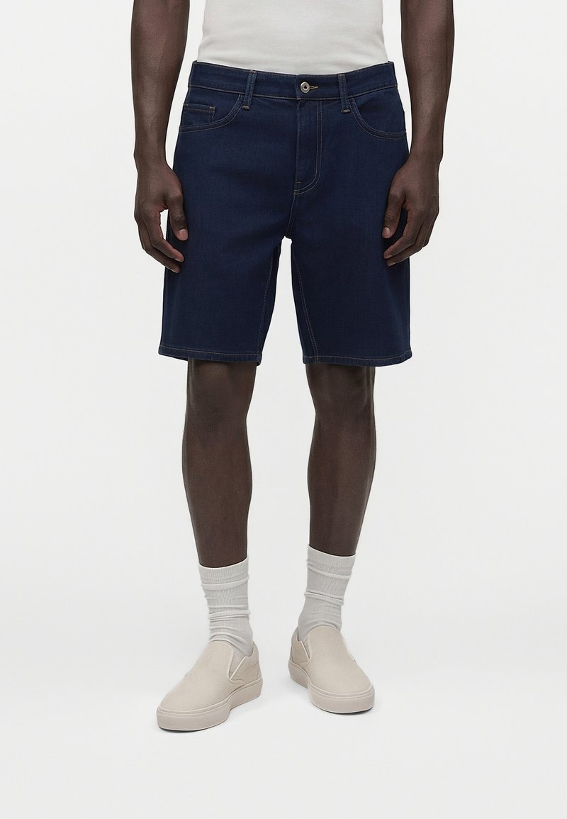 Άνδρας που φοράει σκουρό μπλε τζιν σορτς, λευκές κάλτσες, μπεζ παπούτσια χωρίς κορδόνια και λευκό τοπ, στέκεται μπροστά σε ένα απλό φόντο.