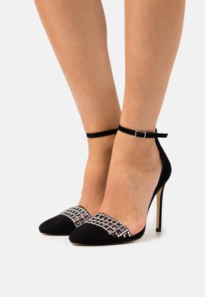 Chaussures à talons hauts noires avec des brides de cheville et des embellissements en strass argentés, portées pieds nus devant un fond uni.