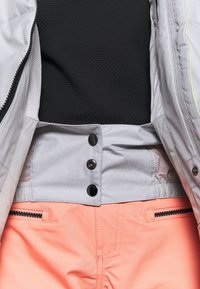 Haut noir rentré dans un pantalon corail avec poches zippées à l'avant, associé à une veste gris clair partiellement ouverte montrant des boutons-pression noirs.