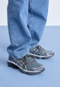 Szürke és zöld sportcipők, hálós felsőrész, fekete részletek, és párnázott talp mintás bordázattal. Világoskék farmernadrággal viselve.