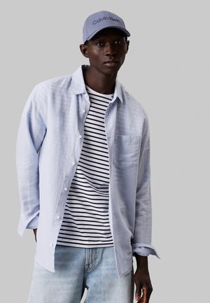 Camisa button-up de color azul claro sobre una camiseta de rayas de manga corta. Se lleva con un denim azul claro y una gorra a juego de color azul con un logotipo bordado.