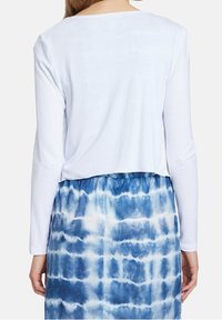 Weißes Langarmoberteil mit einem verkürzten Saum, kombiniert mit einem blauen Batikrock, der verschiedene Blautöne aufweist und einen gerafften elastischen Bund hat.