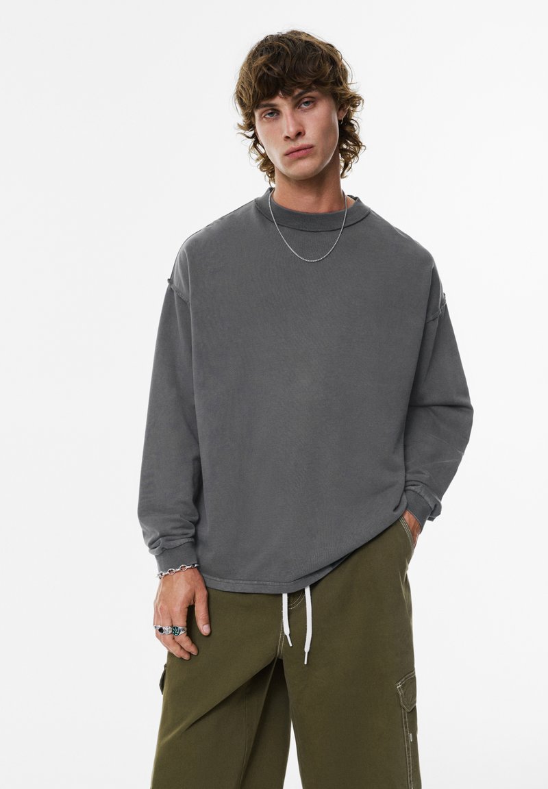 Chemise grise à manches longues avec une coupe décontractée, en tissu texturé et épaules tombantes, associée à un pantalon vert olive avec cordons de serrage et poches.