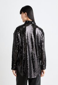 Schwarzes Paillettenhemd mit einer glänzenden, reflektierenden Textur, ausgestattet mit einem hohen Kragen und langen Ärmeln, das für einen oversized Schnitt entworfen ist.