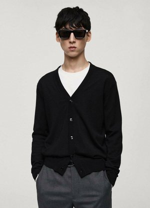 Črn cardigan z V-izrezom in sedmimi gumbi, kombiniran z belim majico. Model nosi črne sončna očala in sivo hlače.