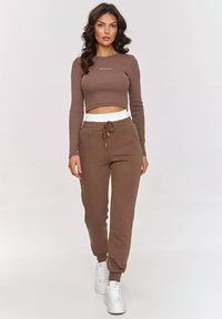 Brązowy prążkowany długi rękaw crop top z logo, w połączeniu z dopasowanymi dresami z elastycznym paskiem w talii i ściągaczami przy kostkach oraz białymi sneakersami.