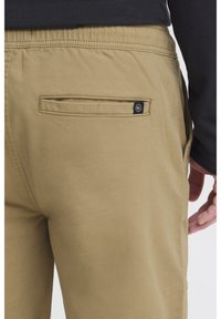 Khaki byxor med elastisk midjeband, med en bakficka som har en knappdetalj. Tyget verkar slätt med minimal textur.