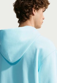 Νεαρός με σγουρά μαλλιά που φοράει ανοιχτό μπλε φούτερ με κουκούλα, με προφίλ και πλάτη προς τα πλάγια, σε απλό φόντο.