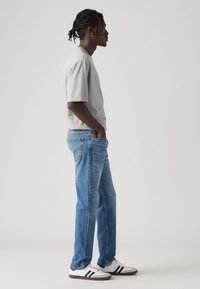 Graues Kurzarm-T-Shirt, blaue Jeans, weiße Sneaker mit schwarzen Streifen, lässig getragen, im Stand-Profil vor einem hellen Hintergrund.