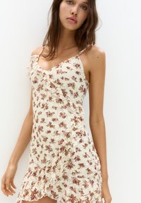 Kräppklänning i krämfärgad med rosa och grönt blommönster, spagettistroppar och volangkant, som visar en genomskinlig textur och delikathet.