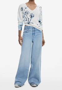 Pull à col en V blanc avec imprimé floral bleu, associé à un jean large bleu clair et des chaussures argentées avec des détails perforés.