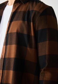 Camisa de cuadros en tonos marrón y negro con diseño de botones, fabricada en un tejido suave con una textura sutil. Camisa blanca visible debajo.