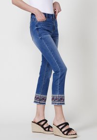 Blå denimjeans med broderad fåll i paisleymönster, rak passform och fransade kanter, kombinerat med svarta kilklacksandaler.