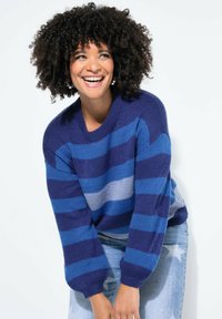 Gestrickter Pullover mit breiten horizontalen Streifen in Marineblau, Blau und Hellgrau. Verfügt über eine lockere Passform und gerippte Bündchen, getragen zu hellblauen Jeans.