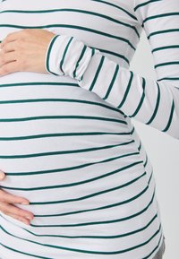 Fehér, sötétzöld vízszintes csíkos, hosszú ujjú felső, amely testhezálló fazonnal és redőzött részletekkel rendelkezik a has körül.