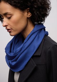 Modrý šátek z měkké tkaniny, volně omotaný kolem krku, přes šedý vršek a černý kabát, s hladkou strukturou a výraznou barvou.