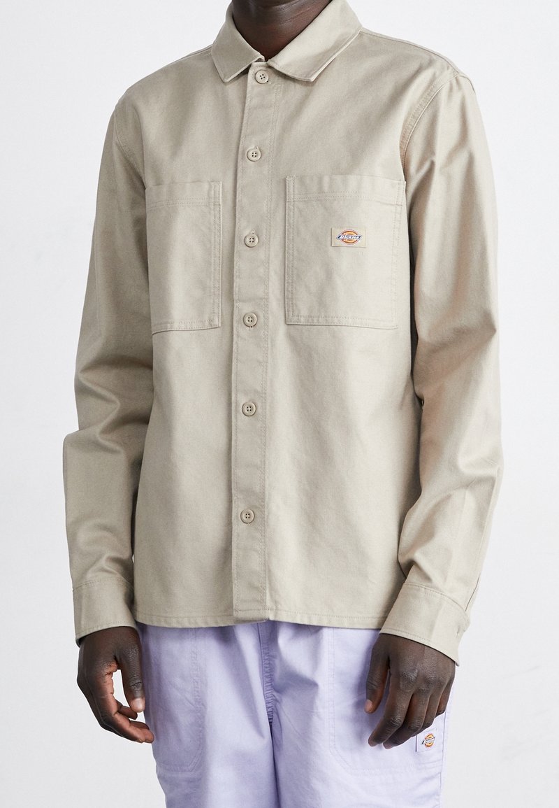 Mężczyzna w beżowej koszuli z guzikami, mającej dwie kieszenie na piersi, oraz w jasnofioletowych spodniach, obie części odzieży z logo marki Dickies.