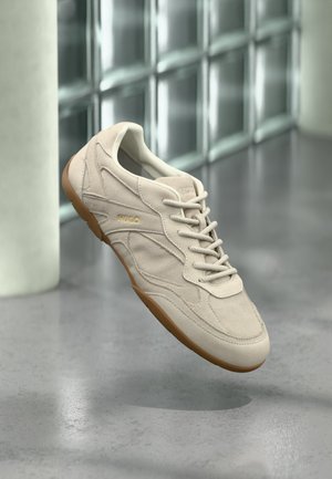Baskets en daim beige avec semelle gomme et lacets, embossées du logo "HUGO", en suspension dans les airs devant un mur en blocs de verre et un sol en béton.