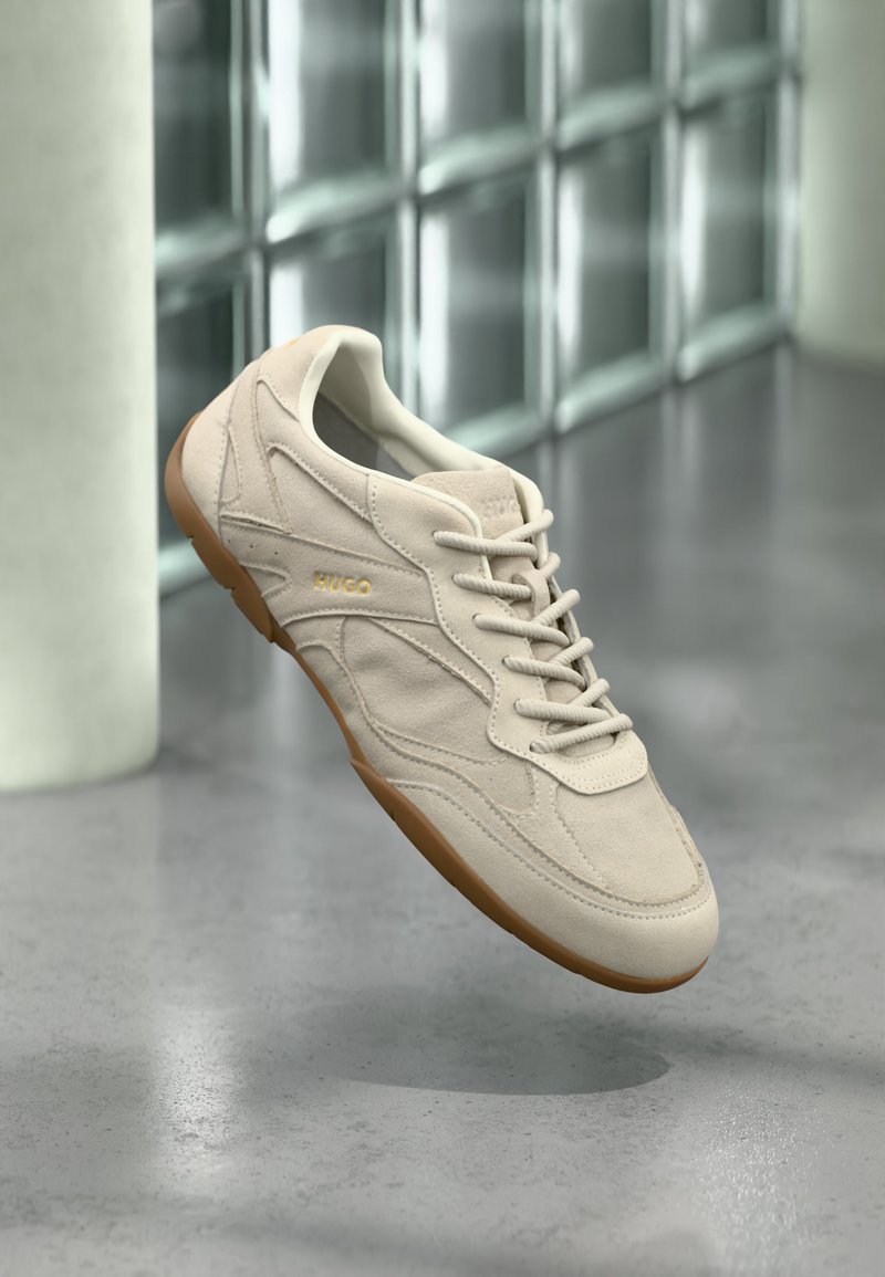 Baskets en daim beige avec semelle gomme et lacets, embossées du logo "HUGO", en suspension dans les airs devant un mur en blocs de verre et un sol en béton.