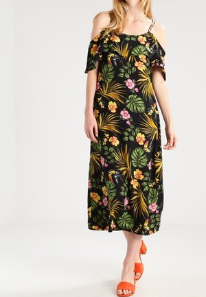 Vrouw draagt een zwarte cold-shoulder jurk met een kleurrijke tropische bloemen- en vogelprint, gecombineerd met feloranje open-toe hakken.