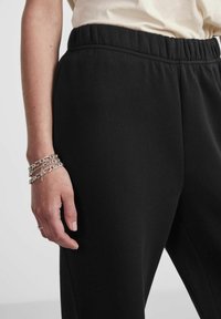 Svarte joggebukser laget av myk stoff, med elastisk midjebånd. En hånd med sølvkjedearmbånd hviler på benet.