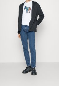 Grå cardigan över en vit t-shirt med en färgglad zebra-grafik, parat med blå jeans och svarta sneakers.