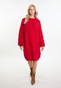 Abito rosso lavorato a maglia con texture a coste, forma oversize e maniche a pompon voluminose. Abbinato a stivali alla caviglia a punta color sabbia su uno sfondo neutro.