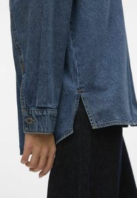 Blue denim shirt with a relaxed fit, side slits at the hem, and buttoned cuff detail. The fabric features a subtle texture and classic stitching.